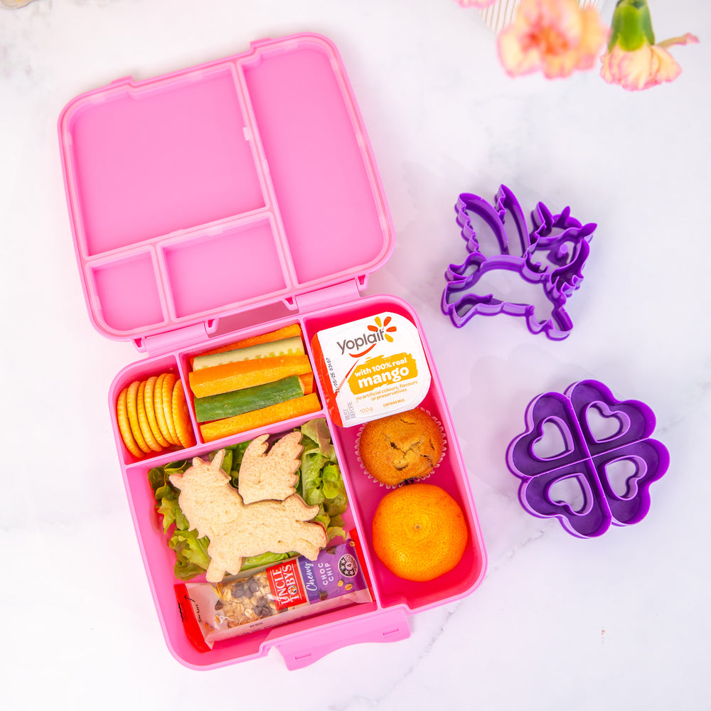 Pink bento box with food compartments on a white background with purple cookie cutters.